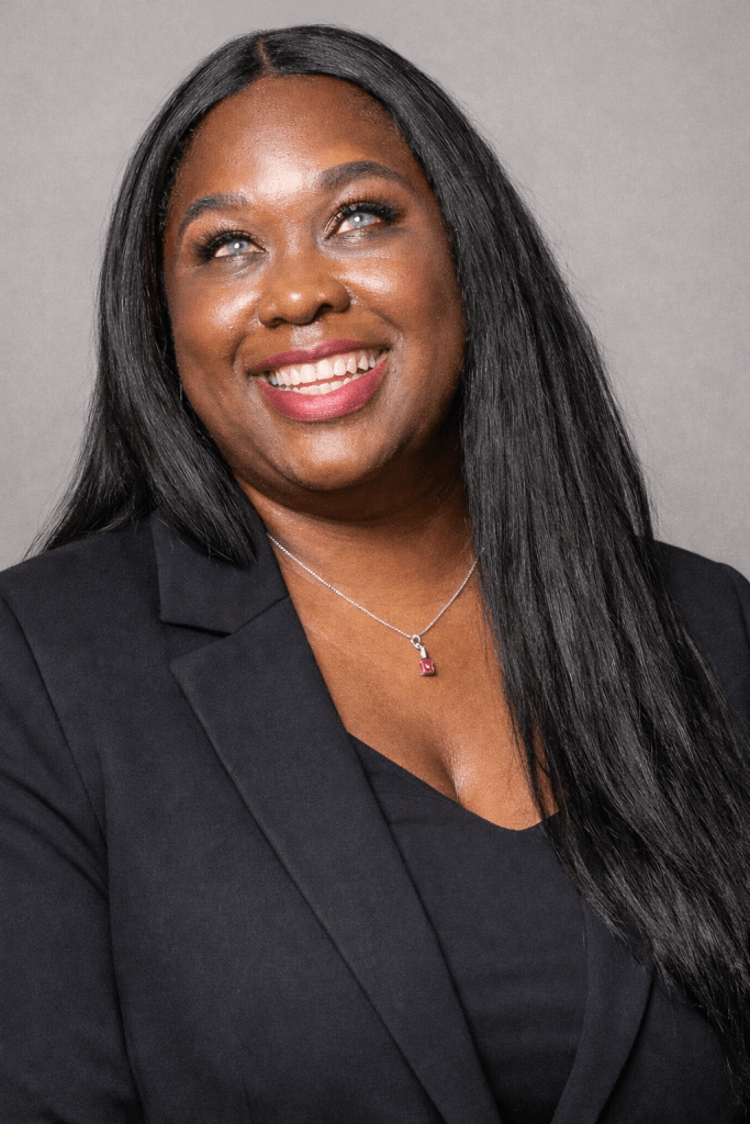 A professional headshot of LaKisha Holmes smiling warmly. She has long straight black hair, is wearing a black blazer and black top, and a necklace with a small pendant. The background is a soft gray studio backdrop.